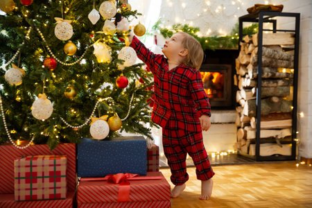 Cute toddler in red plaid pajamas celebrating Christmas at home. Child having fun decorating Christmas tree. Beautiful cozy house with fireplace, wrapped gifts on floor.の写真素材