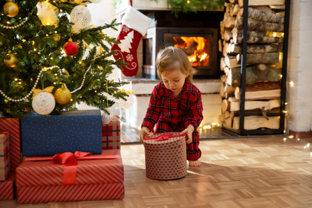 Traditional celebrating Christmas day at home. Cute baby in red plaid pajamas opening gifts at decorated Christmas tree. Beautiful cozy house with fireplace, wrapped giftboxes on floor.の写真素材