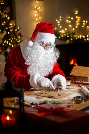 Jolly Santa Claus baking ginger bread, making ginger bread house. Warm, cozy festive room with fireplace, Christmas treeの写真素材