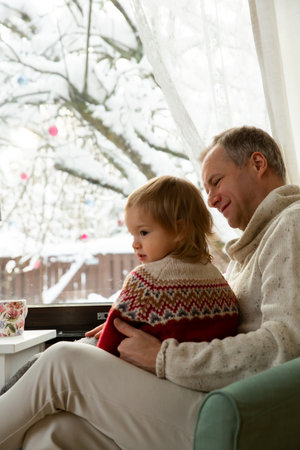 Middle age man and toddler sitting home in a chair by window with with scenic winter view of trees in snow. Bonding and quality family time together.の写真素材
