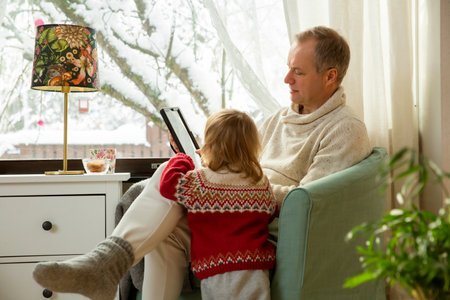 Middle age man and toddler sitting home in a chair by window with with scenic winter view of trees in snow. Family using tablet together.の写真素材