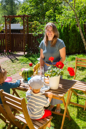 Mother cutting watermelon with toddlers at a backyard table. Sunny summer day, family time, and cozy outdoor moment full of warmth.の写真素材