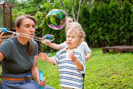 Mother blowing soap bubbles with toddlers in the backyard. Joyful family time outdoors on a sunny, warm day.の写真素材