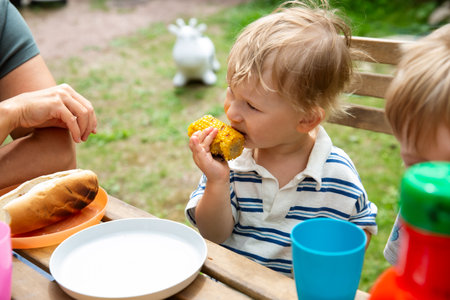 Mother and children eating grilled hot dogs and corn at a backyard table. Cozy family lunch outdoors on a warm summer day.の写真素材