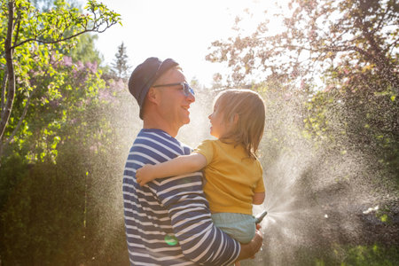 Father holding his laughing happy 2-year-old son while watering plants in the garden on a sunny summer day. Family happiness and outdoor joy.の写真素材