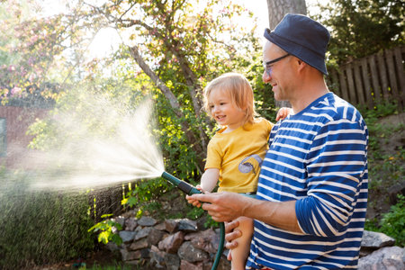 Father holding his laughing happy 2-year-old son while watering plants in the garden on a sunny summer day. Family happiness and outdoor joy.の写真素材