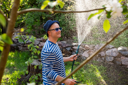 Middle-aged man in a sun hat watering lawn and garden plants with a hose in the backyard on a sunny day. Relaxing gardening activity, summer vibesの写真素材