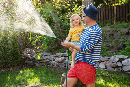 Father holding his laughing happy 2-year-old son while watering plants in the garden on a sunny summer day. Family happiness and outdoor joy.の写真素材