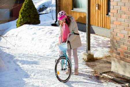 Tween girl in helmet stands next to bicycle, checking her bag near house on a snowy day. Finnish winter landscape, quiet street, school commute sceneの写真素材