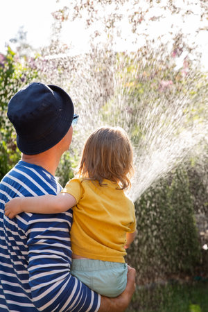 Father holding his laughing happy 2-year-old son while watering plants in the garden on a sunny summer day. Family happiness and outdoor joy.の写真素材