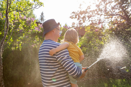 Father holding his laughing happy 2-year-old son while watering plants in the garden on a sunny summer day. Family happiness and outdoor joy.の写真素材