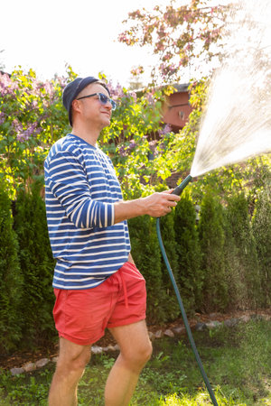 Middle-aged man in a sun hat watering lawn and garden plants with a hose in the backyard on a sunny day. Relaxing gardening activity, summer vibesの写真素材