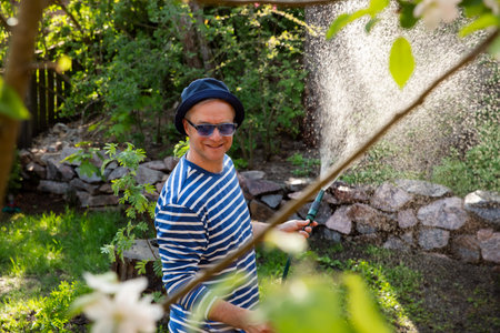 Middle-aged man in a sun hat watering lawn and garden plants with a hose in the backyard on a sunny day. Relaxing gardening activity, summer vibesの写真素材