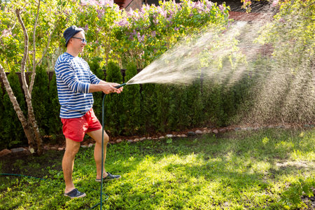 Middle-aged man in a sun hat watering lawn and garden plants with a hose in the backyard on a sunny...