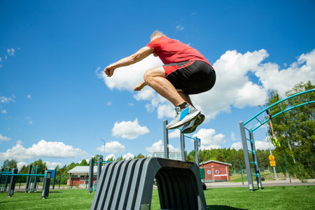 Middle-aged man performs a box jump on a plyo box in a sunny outdoor gym, showcasing explosive strength and balance training.の写真素材