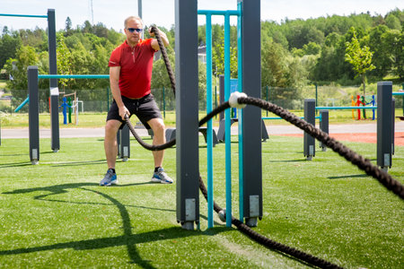 Active middle-aged man training with battle ropes at an outdoor gym on a sunny summer day. Fitness, strength, and healthy lifestyle in the open airの写真素材