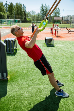 Middle-aged man performs bodyweight ring rows on gymnastic rings in an outdoor fitness park....