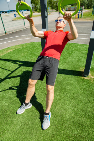 Middle-aged man performs bodyweight ring rows on gymnastic rings in an outdoor fitness park....
