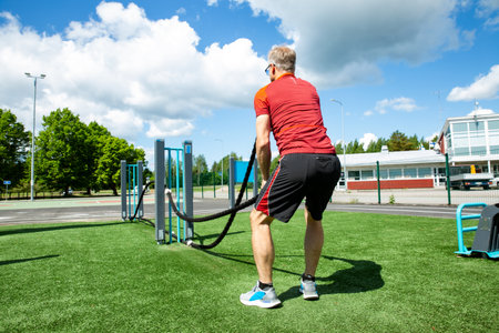 Active middle-aged man training with battle ropes at an outdoor gym on a sunny summer day. Fitness,...