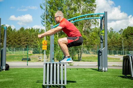 Middle-aged man performs a box jump on a plyo box in a sunny outdoor gym, showcasing explosive strength and balance training.の写真素材