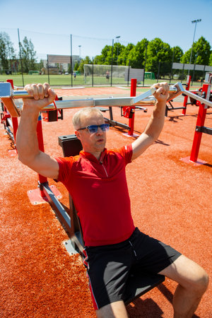 Middle-aged man using outdoor gym chest press machine to train shoulders and triceps. Focused...
