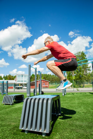 Middle-aged man performs a box jump on a plyo box in a sunny outdoor gym, showcasing explosive strength and balance training.の写真素材