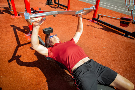 Middle-aged man performing bench press outdoors. Focus on chest, shoulders, triceps. Maintaining healthy lifestyle through regular strength training.の写真素材