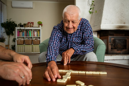A grown son and his elderly father enjoy a quiet game of dominoes in the living room, sharing...