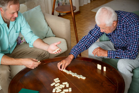 A grown son and his elderly father enjoy a quiet game of dominoes in the living room, sharing...