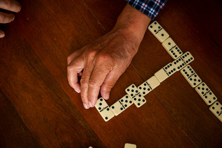 Close-up of adult son and elderly fathers hands as they play dominoes together at home, highlighting...