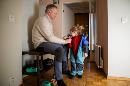 Father helping toddler get dressed in overall at hallway, child learning to put on clothes before...