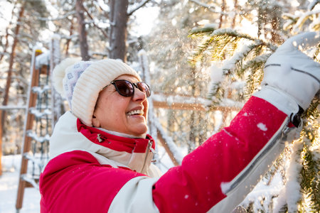 Smiling mature woman shaking snow from branches in golden sunlight, celebrating winter, menopause vitality and active lifestyle outdoors in Finland.の写真素材