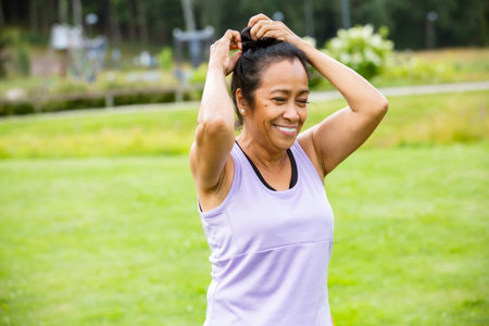 Confident middle aged woman of color tying hair for outdoor yoga in the park, promoting wellness, mindfulness, fitness, and active lifestyle.の写真素材