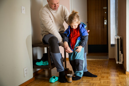 Toddler zipping overall by himself in hallway, father nearby supporting child getting dressed for cold autumn outdoor walk.の写真素材