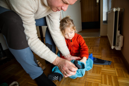 Father helping toddler get dressed in overall at hallway, child learning to put on clothes before going outside in cold season.の写真素材