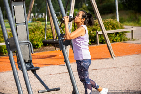 Confident Asian woman drinking water after outdoor workout in sunny morning park, embracing fitness,...