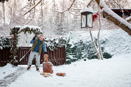 Middle-aged man chopping firewood in snowy backyard on a cold winter day, warm clothing, lifestyle...