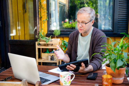 Middle aged man working remotely on laptop at cozy sunny patio, digital nomad lifestyle, outdoor home office, freelance and productivity conceptの写真素材