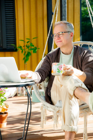 Middle-aged man working remotely from laptop and drinking coffee in hanging chair on sunny patio, relaxed lifestyle and modern home office.の写真素材