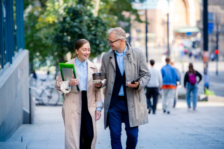 Successful business man and woman walking in city street, smiling and talking with laptop and coffee. Modern urban teamwork and success concept.の写真素材