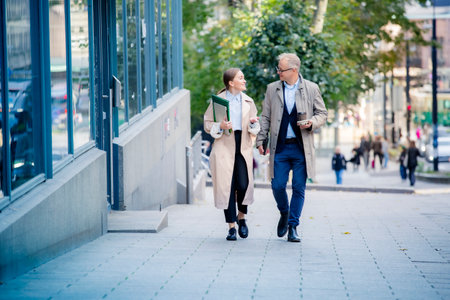 Successful business man and woman walking in city street, smiling and talking with laptop and coffee. Modern urban teamwork and success concept.の写真素材