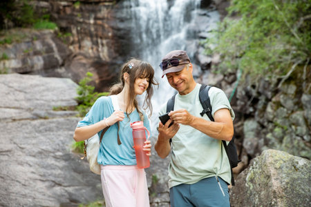 Father and teenage daughter checking smartphone during mountain hike by waterfall in Norway. Family travel, GPS navigation and outdoor adventure.の写真素材