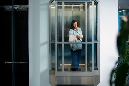 Professional businesswoman using smartphone inside modern glass elevator. Corporate communication,...