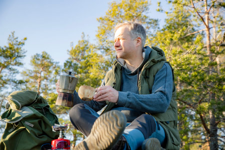 Male hiker brewing coffee on rocky cliff in Finland, overlooking forest. Scandinavian adventure travel and outdoor lifestyle.の写真素材