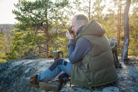 Male hiker taking photos with vintage camera on scenic cliff in Finland. Travel photography, adventure tourism, outdoor lifestyleの写真素材