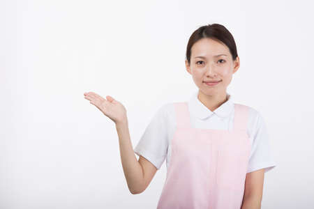 A young woman in a white coat and pink apron smiling as she raises her hand in front of a white background.の写真素材