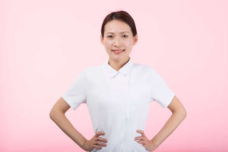 A young woman in a white coat smiling with her hands on her hips in front of a pink background.の写真素材