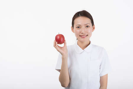 A young woman in a white coat smiling with an apple in her hand in front of a white background.の写真素材