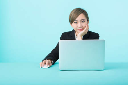 A young woman in a black suit smiling as she operates her laptop with a green desk.の写真素材