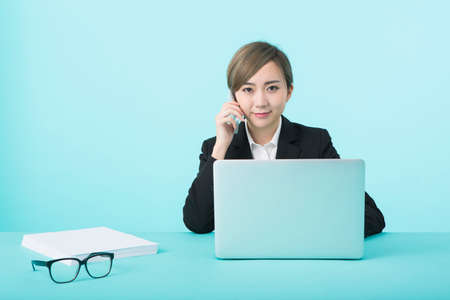 A young woman in a black suit smiling with her smartphone in her ear while operating a computer on a green desk.の写真素材